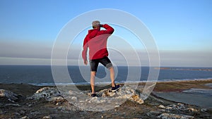 A slender man in a red jacket stands on a high mountain in a strong wind and watches a flying bird in the sky against