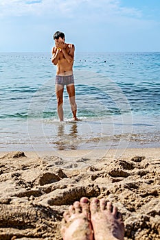 A slender man wipes his wet face with his hands when he comes out of the sea on the beach. Blurry female feet soiled in the sand