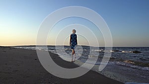 Slender sweet woman in a dress walks barefoot on a sandy beach against the background of running waves and the sea on a