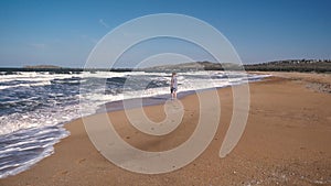 Slender sweet woman in a dress walks barefoot on a sandy beach against the background of running waves and the sea on a