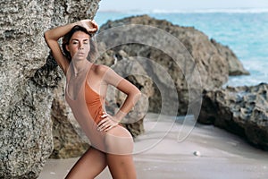 Slender tanned girl in swimsuit posing on beach with sand and large stones