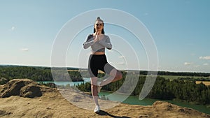 slender woman in gray top and black sports shorts practices yoga outdoors in nature. Pretty lady raises her hands up in