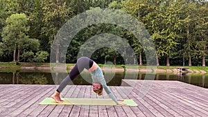 A slender woman lie on mat on wooden platform by pond, does flow yoga in park