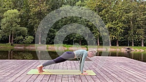 A slender woman lie on mat on wooden platform by pond, does yoga in park
