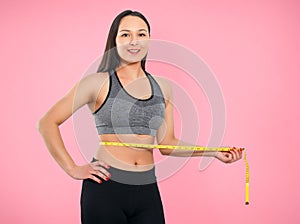 A slender woman measures her waist with a measuring tape. n a pink background