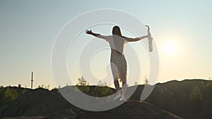 slender woman in white dress stands on top of mountain, hands to the side, holding saxophone in one hand against