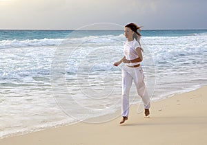 Slender woman in a white tracksuit jogging along the edge of the waves on the sandy beach of the sea