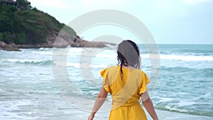A slender young beautiful brunette girl in a yellow dress enjoys the wind breeze while walking along the beach by the bay.