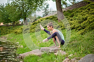 Slender young brunette yogi performs challenging yoga exercises on the green grass in the summer against the backdrop of nature
