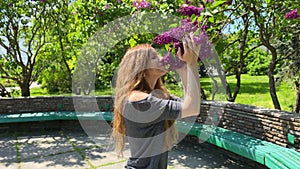 A slender young girl inhales the aroma of spring lilac flowers in the park.