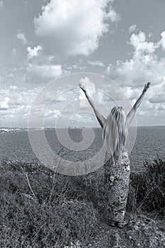 Slender young girl with long blonde hair on the mountain by the seashore with hands up to the sky. People connecting with nature