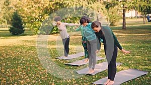 Slender young ladies are practising yoga outdoors in park under guidance of experienced teacher, woman is speaking and