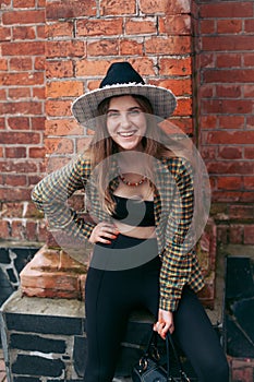 A slender young smiling girl with blond long hair, in black leggings, a tank top, a beige jacket and a hat