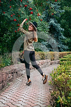 A slender young smiling girl with blond long hair, in black leggings, a tank top, a beige jacket and a hat