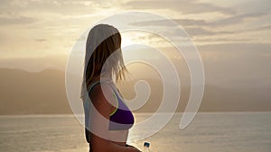 slender young woman in purple swimsuit with bottle of water in her hands turns to camera with beach and sea or ocean in