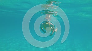 A slender young woman swims in a scuba mask and snorkel underwater in a transparent sea with a sandy bottom