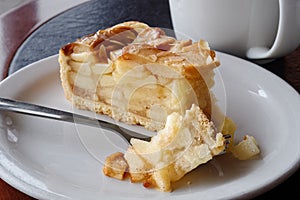 A slice of apple pie with pie on fork on white ceramic plate next to a fork. Black and brown table.