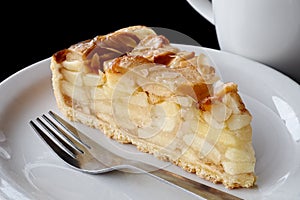 A slice of apple pie on white ceramic plate with fork. Black background.