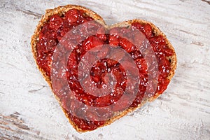 Slice of bread in shape of heart with strawberry jam for breakfast. Rustic background