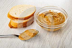 Slice of bread, spoon with peanut butter, transparent bowl with peanut butter on wooden table