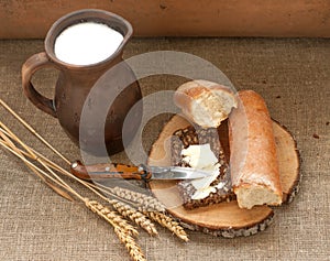 A slice of buttered bread on a wooden board, a crock of milk and mature ears