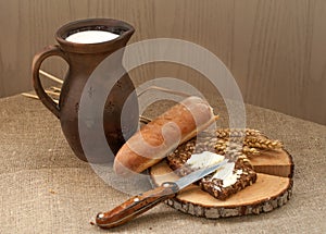 A slice of buttered bread on a wooden board, a crock of milk and mature ears