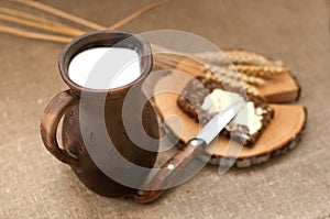 A slice of buttered bread on a wooden board, a crock of milk and mature ears