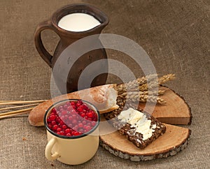 A slice of buttered bread on a wooden board, a crock of milk, a metal cup of red currants and mature ears