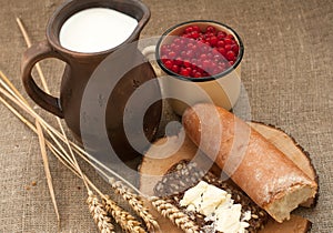 A slice of buttered bread on a wooden board, a crock of milk, a metal cup of red currants and mature ears