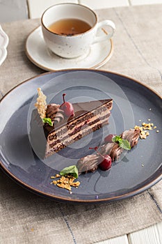 Slice of chocolate cake with fresh cherries on a plate and cup of tea on the table