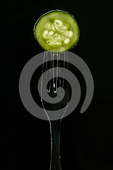 Slice of fresh green cucumber on fork on dark black background. silhouette of a fork in the back light