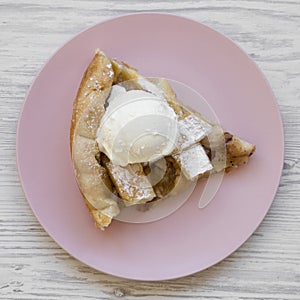 Slice of homemade apple pie with ice cream on pink plate over white wooden surface, overhead view. Flat lay