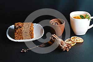 Slice of layered honey cake decorated with anise star, dessert fork, mint, dried lemons, sticks of cinnamon, raw eggs, cocoa, cup