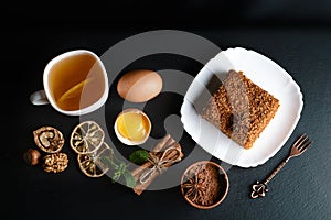 Slice of layered honey cake decorated with anise star, dessert fork, mint, dried lemons, sticks of cinnamon, raw eggs, cocoa, nuts