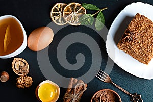 Slice of layered honey cake decorated with anise star, dessert fork, mint, dried lemons, sticks of cinnamon, raw eggs, cocoa, nuts