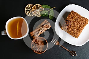 Slice of layered honey cake decorated with anise star, dessert fork, mint, dried lemons, sticks of cinnamon, raw eggs, cocoa, nuts