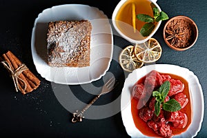 Slice of layered honey cake decorated with anise star, dessert fork, mint, dried lemons, sticks of cinnamon, strawberry jam, cocoa