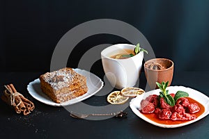 Slice of layered honey cake decorated with anise star, dessert fork, mint, dried lemons, sticks of cinnamon, strawberry jam, cocoa