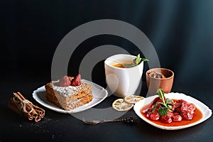 Slice of layered honey cake decorated with anise star, dessert fork, mint, dried lemons, sticks of cinnamon, strawberry jam, cocoa