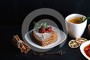 Slice of layered honey cake decorated with strawberry, dessert fork, mint, dried lemons, sticks of cinnamon, strawberry jam, cup o