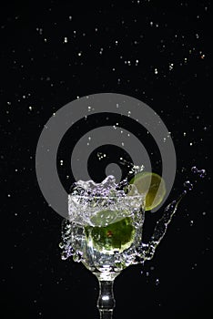 a slice of lemon falling into a glass cup with clear water causing splashes. Isolated on dark background