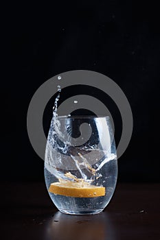 a slice of lemon falls into water in clear glass, splashes fly, citrus in glass with drink, dark background, close-up