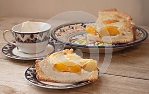 Slice of peach apricot pie or cheese sour cream cake on bright saucer with tea cup on wooden table