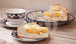 Slice of peach apricot pie or cheese sour cream cake on bright saucer with tea cup on wooden table