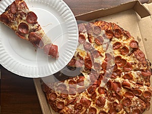 Slice of pepperoni pizza on a paper plate next to a large delivered pizza.
