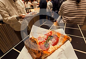 Slice of pizza with with cheese and greens, on table of small pizzeria with crowd of customers, Italy