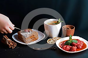 Slice of poured honey cake, cook decorated with strawberries, dessert fork, mint, dried lemons, sticks of cinnamon, strawberry jam