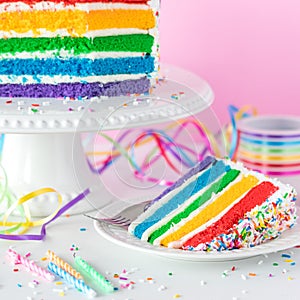 A slice of rainbow birthday cake with the remaining cake on a stand in behind.