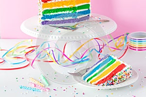 A slice of rainbow birthday cake with the rest of the cake on a stand in behind.