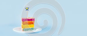 Slice of Rainbow cake with birning candle in the shape of star on white round plate isolated on blue background. Happy bithday,
